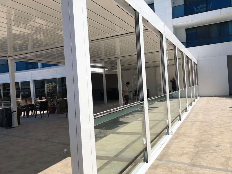 Otel dış alanına kurulan beyaz doğramalı giyotin cam koridoru; camlar kapalı, içeride masa düzeni hazırlanıyor