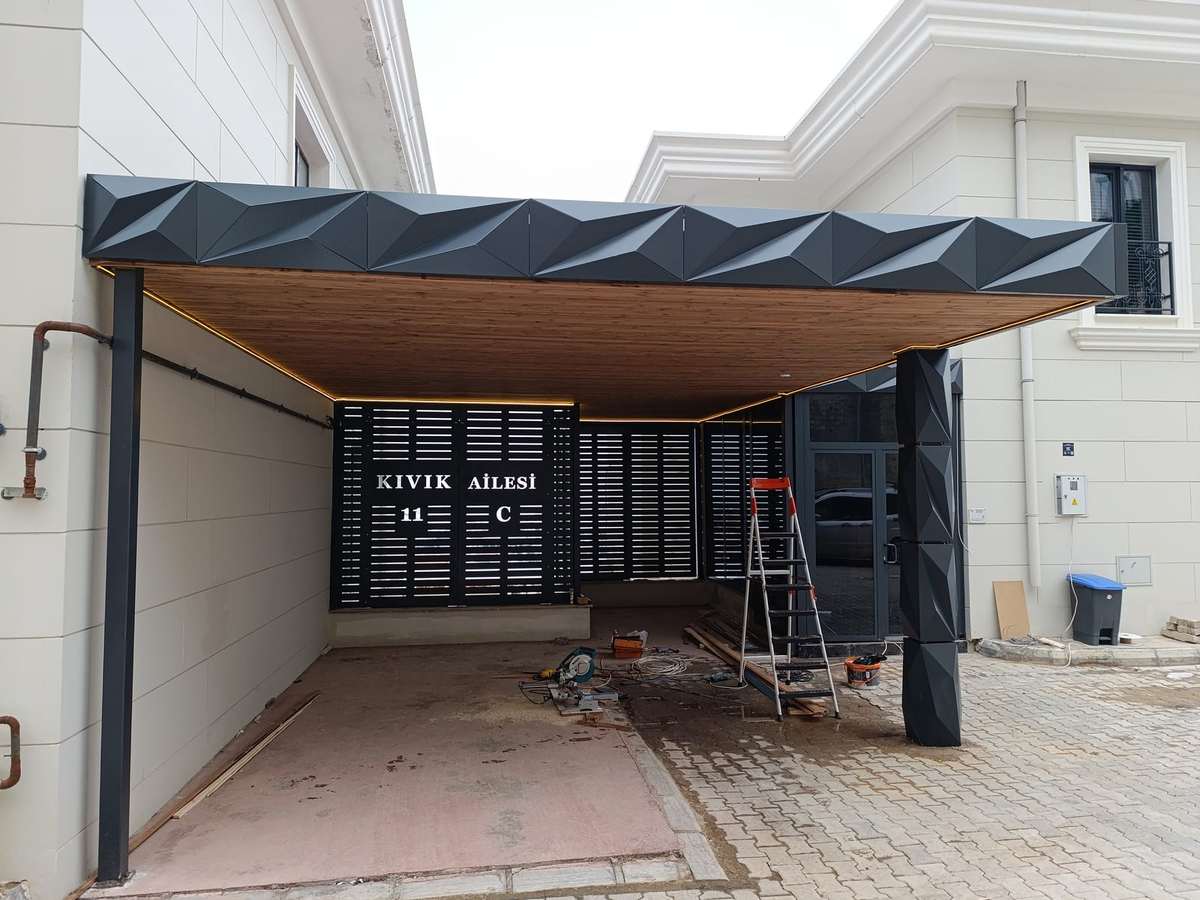 Villa girişine kurulan araç gölgeliği; alınlıkta üç boyutlu kırılımlı antrasit kompozit panel, tavanda ahşap desenli kaplama ve gömme LED şerit, arkada lazer kesim desenli bahçe kapısı